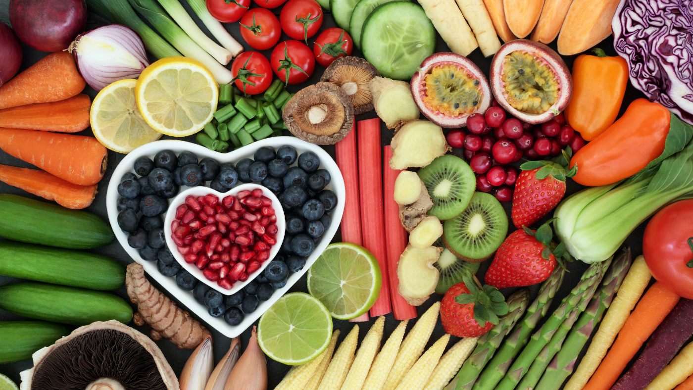 Assortiment coloré de fruits et légumes frais incluant carottes, tomates, baies, agrumes et légumes verts. Un bol en forme de cœur au centre contient des myrtilles et des graines de grenade.