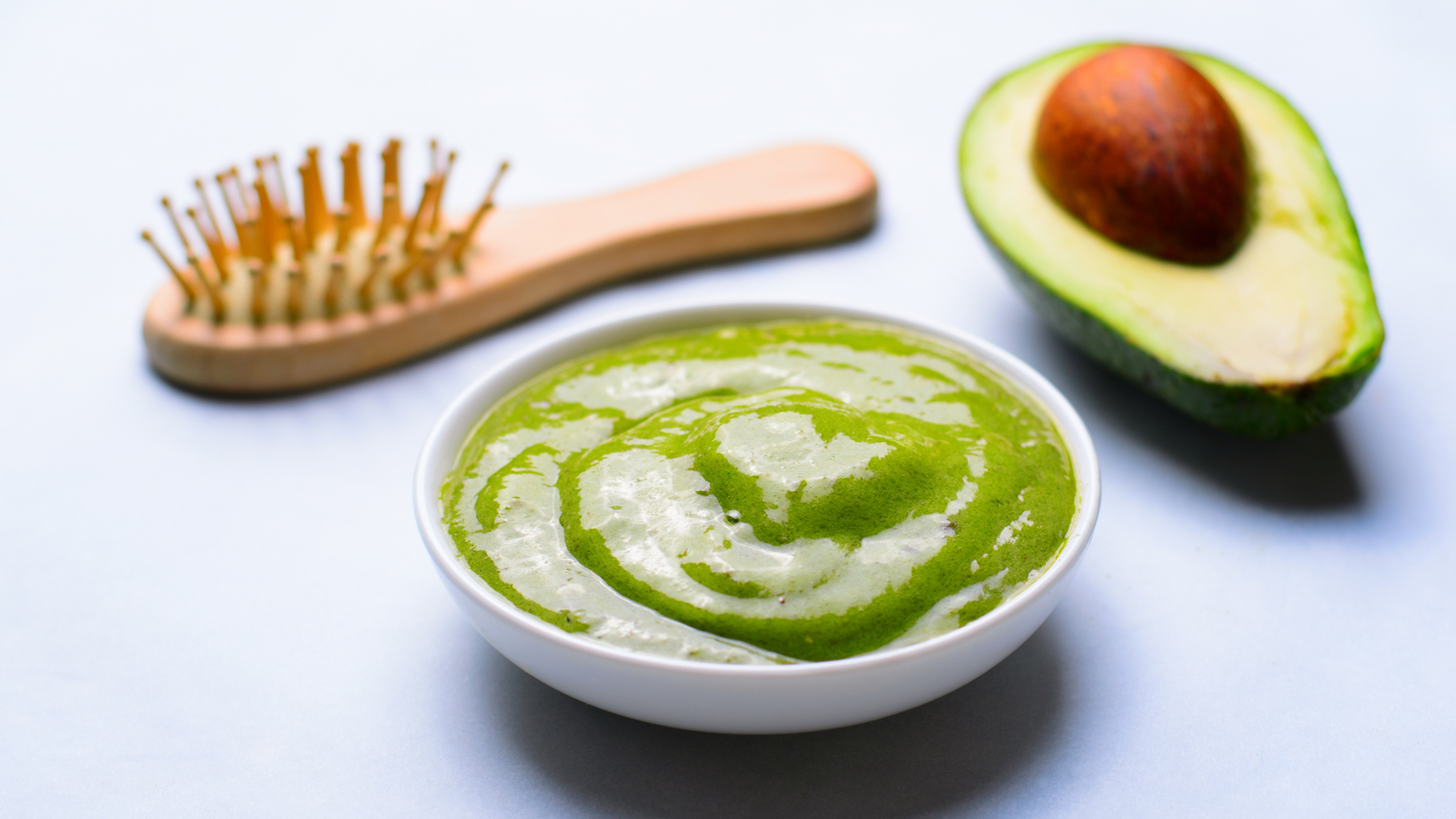 Image d'une brosse à cheveux en bois, d'un bol de masque capillaire à l'avocat vert et d'un avocat coupé en deux, illustrant des produits de soins capillaires naturels.