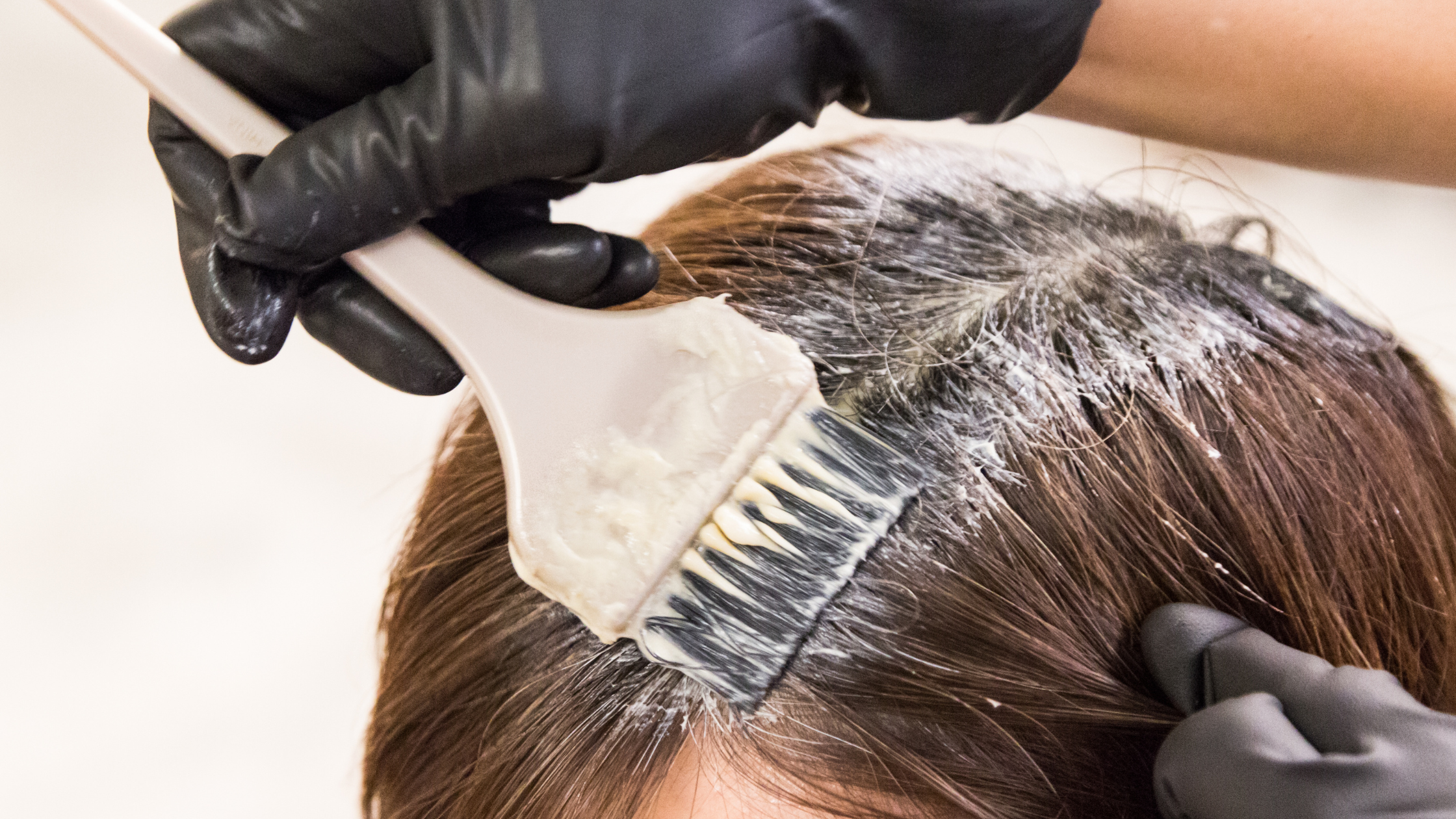 L'image montre une personne appliquant une coloration capillaire avec précision dans un salon, utilisant des gants et un pinceau pour couvrir les racines des cheveux, illustrant ainsi une technique professionnelle de coloration.